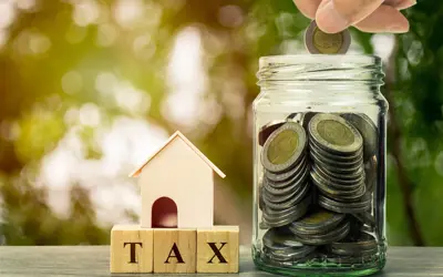 A small wooden model house, beside a jar of money coins, and a hand putting a coin in the jar.